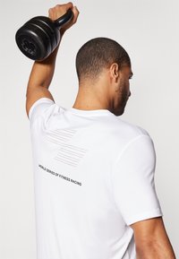 Hvit treningst skjorte med korte ermer som har et grafisk design på ryggen. Teksten sier "WORLD SERIES OF FITNESS RACING." En svart kettlebell hevet over hodet.