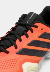 Κοντινό πλάνο αθλητικού παπουτσιού σε πορτοκαλί και μαύρο χρώμα με μαύρα κορδόνια, τρεις σκούρες γκρι πλευρικές ρίγες και παχύ, λευκό πάτο.