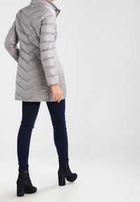 Grå polstret frakke med et chevronmønster, høj krave og lange ærmer, parret med marineblå leggings og sorte høje ankelstøvler.
