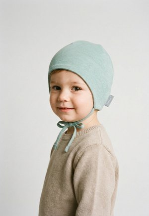 Kleinkind trägt eine hellgrüne Haube, die unter dem Kinn gebunden ist, und einen beigen Pullover, lächelt leicht vor einem einfarbigen hellen Hintergrund.