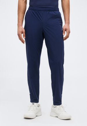 Férfi sötétkék, elasztikus derékpántú sportnadrágban és fehér sportcipőben, világos, egyszínű háttér előtt állva.