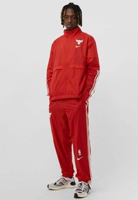Tuta rossa realizzata in materiale leggero, con giacca zip-up decorata con un logo e pantaloni coordinati con strisce laterali e loghi.