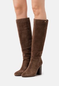 Bottes hauteur genou en suède marron avec un talon carré, présentant une texture lisse et un revers replié, accentuées par un petit emblème doré.