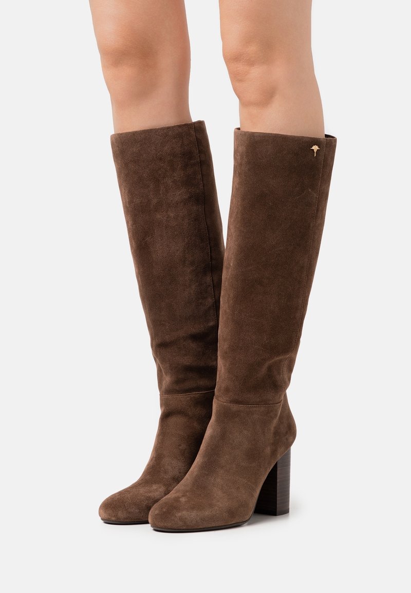 Bottes hauteur genou en suède marron avec un talon carré, présentant une texture lisse et un revers replié, accentuées par un petit emblème doré.