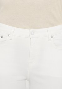 Fehér farmernadrág sima textúrával, elülső gombos záródással, övön lévő hurkokkal és két elülső zsebbel.