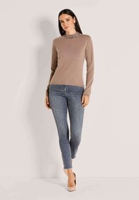 Vrouw staat in een beige lange mouwen trui, een gestreepte coltrui, grijze skinny jeans en nude hakken tegen een effen lichte achtergrond.