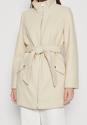 Vrouw draagt een beige wollen jas met een hoge kraag, rits aan de voorkant, ceintuur in de taille en klepzakken met knopen.