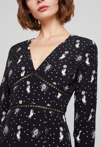 Černé šaty s dlouhými rukávy, zdobené bílými květinovými a rukodělnými vzory. V- výstřih s krajkovými detailemi kolem pasu a korpusu. Jemná tkanina.