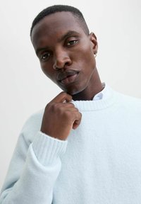 Lys blå sweater med ribbet halsudskæring og manchetter, lavet af blødt materiale. Nærbillede af en persons hånd nær hagen, der viser teksturen.