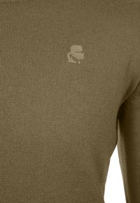 Olivově zelený pletený svetr s texturovaným povrchem a malým, vyraženým logem na hrudi. Jednobarevný, bez vzorů nebo zdobení.