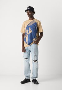 Beige T-skjorte med blå grafisk design, lyseblå jeans med slitasjedetaljer og svarte sko. Svart caps med hvit logo.