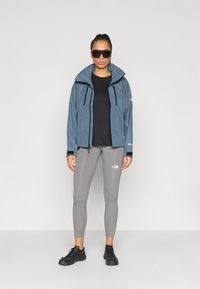 Αδιάβροχο μπλε μπουφάν με ψηλό γιακά, μαύρο τοπ, γκρι κολάν και μαύρα παπούτσια. Διαθέτει ρυθμιζόμενους φερμουάρ και διακριτικές λεπτομέρειες με λογότυπο.