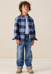 Blauw en marineblauw geruite button-up shirt met een bruine corduroy kraag, gecombineerd met lichtblauwe jeans en beige schoenen, staande op een neutrale achtergrond.