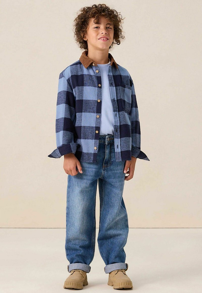 Blauw en marineblauw geruite button-up shirt met een bruine corduroy kraag, gecombineerd met lichtblauwe jeans en beige schoenen, staande op een neutrale achtergrond.