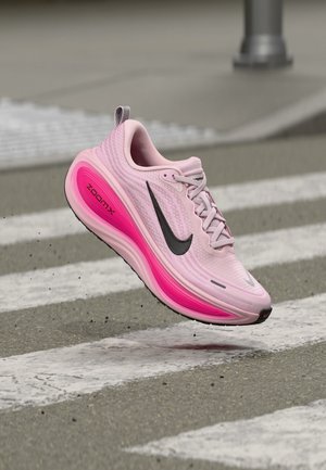Ροζ Nike ZoomX παπούτσι τρεξίματος με μαύρο σήμα, στον αέρα πάνω από διάβαση πεζών, που δείχνει λεπτομέρειες ρόζ σόλας.