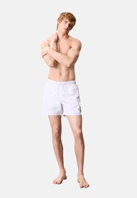 Pantaloni da bagno bianchi con vita elastica e cordoncino, caratterizzati da un piccolo logo nero sulla gamba. Tessuto morbido, lunghezza sopra il ginocchio.
