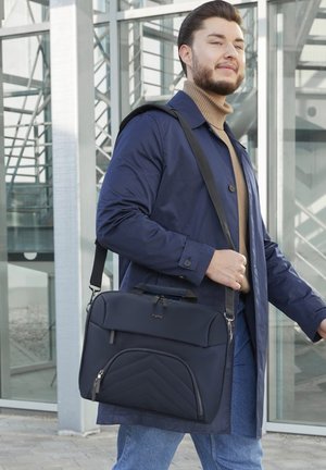 Junger Mann im blauen Mantel, der eine schwarze Laptop-Tasche mit Schultergurt trägt, geht in der Nähe des Eingangs eines Glasgebäudes.