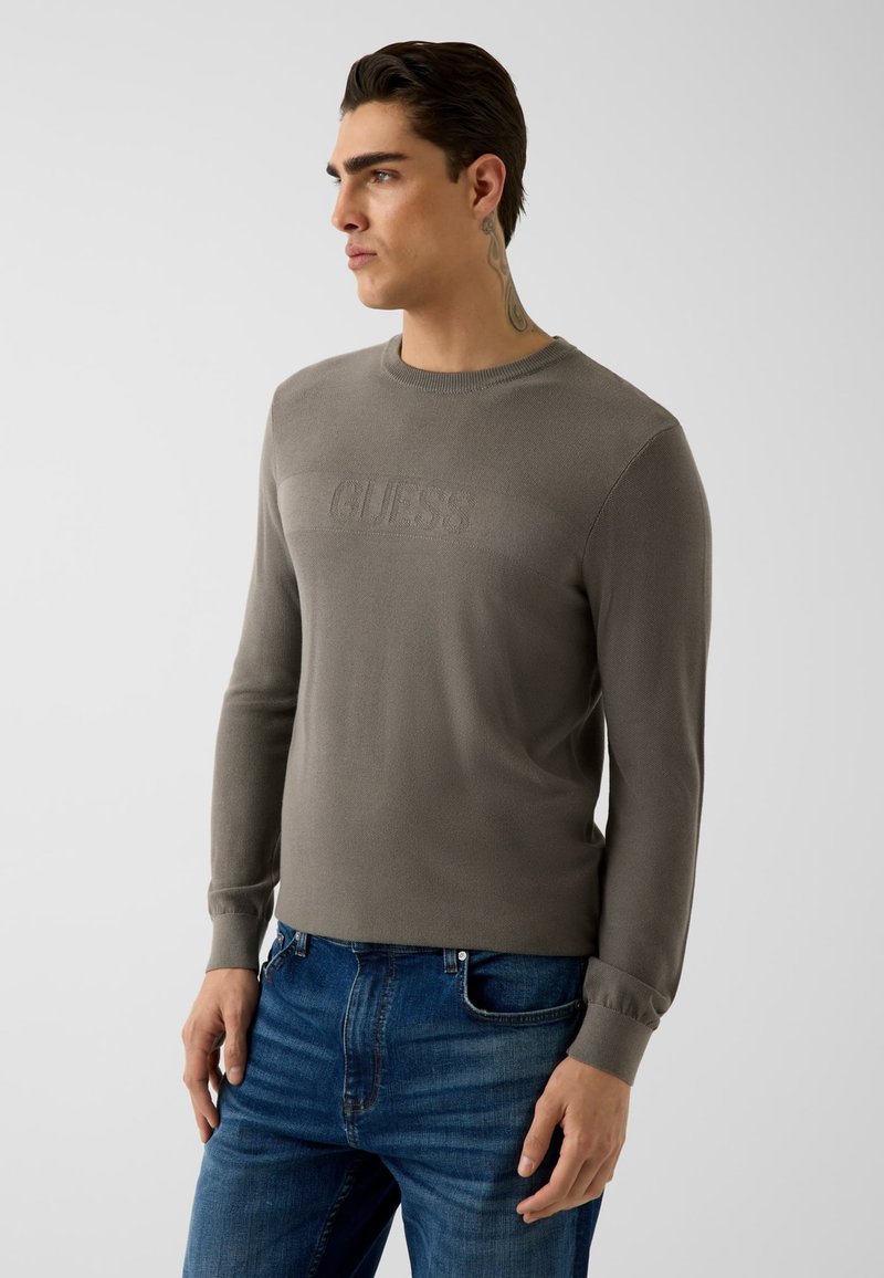 Homme aux cheveux foncés coiffés en arrière et tatouage sur le cou portant un pull GUESS gris à manches longues et un jean bleu, debout sur un fond uni.