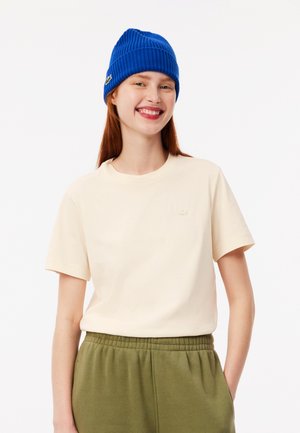 T-shirt crema con maniche corte e un patch logo testurizzato, abbinato a pantaloni della tuta verde oliva e un berretto a coste blu brillante.
