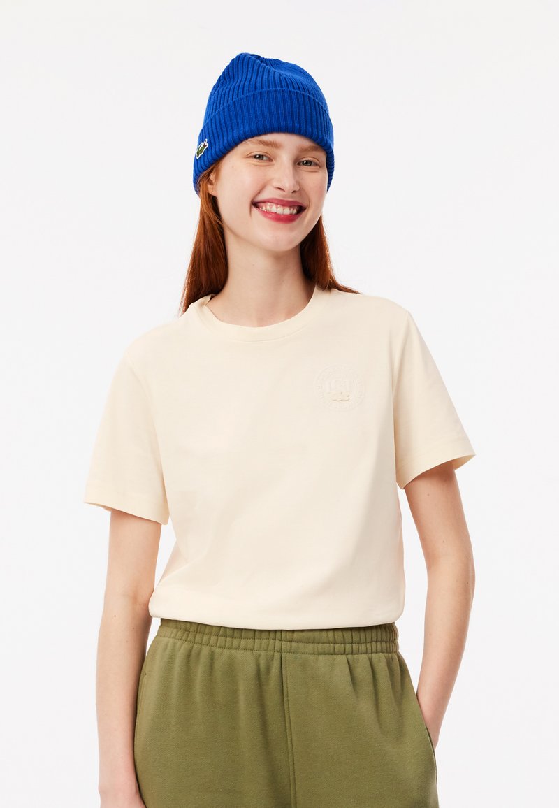 Camiseta crema de manga corta con un parche de logo texturizado, combinada con pantalones de chándal verde oliva y un gorro de canalé azul brillante.