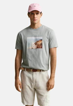 Junger Mann mit rosa Mütze, grauem T-Shirt mit Küstenklippen-Motiv, beigem Gürtel und hellen Shorts, stehend vor einfarbigem Hintergrund.