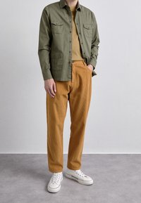 Camisa de botón verde oliva con dos bolsillos en el pecho, camiseta mostaza debajo y pantalones de pana naranja. Zapatillas blancas completan el conjunto.
