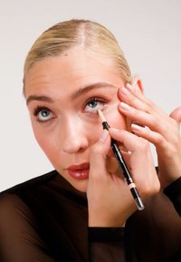 Une personne applique un crayon eyeliner nude à boîtier noir sur la ligne inférieure des yeux, mettant en valeur une peau lisse et des lèvres brillantes.