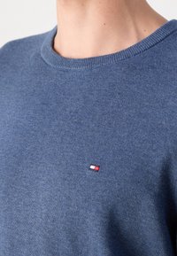 Gros plan d'une personne portant un pull en maille bleu avec un petit logo brodé rouge, blanc et marine sur la poitrine.