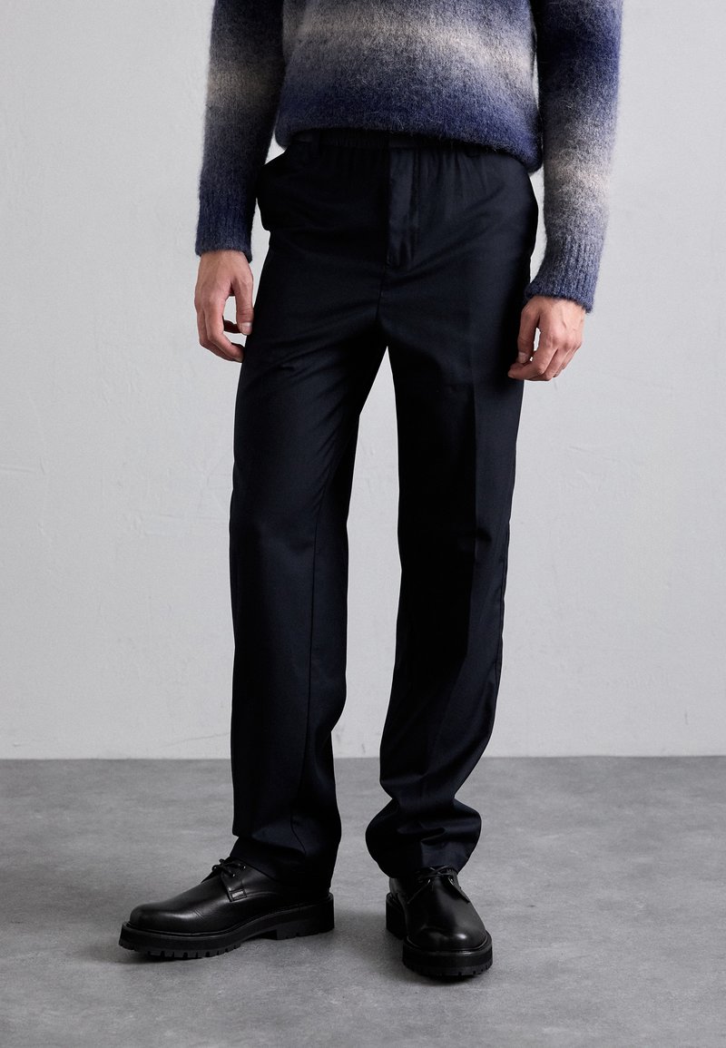 Pantalon noir, ajusté avec une texture lisse et une coupe décontractée. Détail avec une taille subtile et associé à des chaussures en cuir noires.