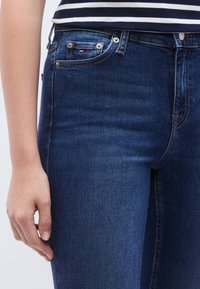 Sötétkék farmernadrág sima textúrával, elülső zsebekkel, fémszerelvényekkel és egy kis, piros-fehér-kék márkajelzésű címkével.