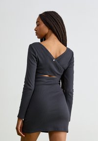 Robe côtelée gris foncé à manches longues, avec un dos en V et des découpes. Présente un détail de bouton à l'arrière. Texture lisse.