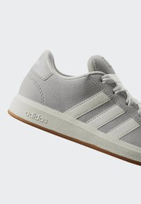 Světlé šedé tenisky Adidas s bílými pruhy, bílými tkaničkami, strukturovanou podrážkou a hnědou podrážkou zespodu, zobrazené z boku na jednobarevném pozadí.