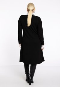 Zwarte jurk met een aangesloten silhouet, lange mouwen en een witte kanten kraagdetail. Het model draagt zwarte kniehoog laarzen.