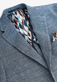Veste bleue tissée avec un tissu texturé. Intérieur doté d'une doublure multicolore à motif de vagues et d'un porte-pochette.
