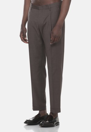 Uomo che indossa pantaloni marroni su misura e mocassini in pelle neri, in piedi contro uno sfondo neutro, ritagliato in vita.