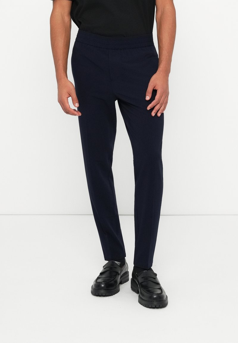 Homme portant un pantalon slim bleu marine et des mocassins en cuir noir, debout devant un fond blanc.