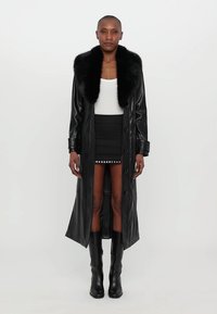 Μαύρο δερμάτινο παλτό με μεγάλο απομίμηση γούνες γιακά, μακριά μάκρος, κλείσιμο με κουμπί και δομημένα μανίκια, συνδυασμένο με κοντή μαύρη φούστα και ψηλές μπότες μέχρι το γόνατο.