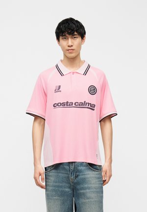 Junger Mann, der ein hellrosa Poloshirt mit schwarzem Besatz, Logos und dem Schriftzug "costa calma" trägt, kombiniert mit ausgewaschenen blauen Jeans.