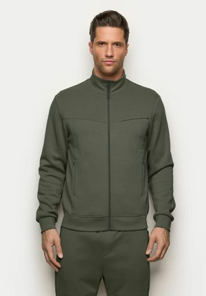 Hombre de pie contra un fondo liso, vestido con chaqueta de cremallera y pantalones a juego de color verde oliva, mirando hacia adelante con expresión neutral.