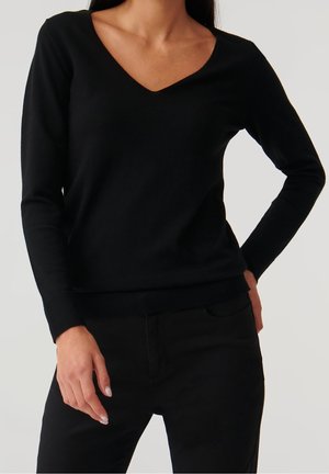 Frau trägt ein eng anliegendes schwarzes Langarmshirt mit V-Ausschnitt und schwarze Hose, eine Hand auf der Hüfte ruhend und die andere seitlich am Körper.