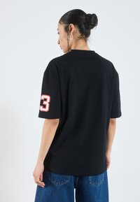Svart bomulls t-shirt med oversized passform, korta ärmar och vit-röd siffra "3" på vänster ärm. Slät textur och rundad halsringning.