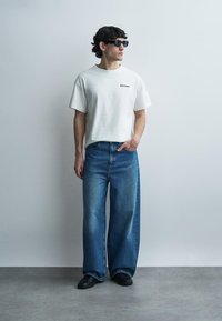 Čovjek koji nosi crne sunčane naočale, bijelu majicu s grafičkim uzorkom, široke plave traperice i crne cipele stoji nasuprot svijetlom zidu.