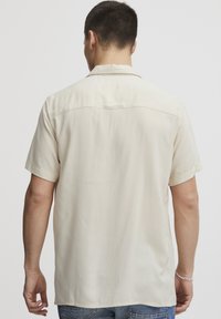 Lys beige kortermet skjorte med omslagskrage, myk tekstur og rett nederkant. Har en søm over ryggbåret og ingen mønstre.