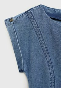 Gulerul cămășii din denim are un singur nasture maro, evidențiind țesătura albastră, cu o cusătură contrastantă și un design de manșetă relaxat.
