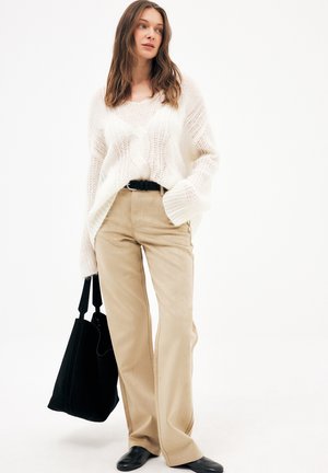Kobieta ubrana w luźny biały sweter z dzianiny, beżowe spodnie z wysokim stanem, czarny pasek, czarne buty i niosąca dużą czarną torbę typu tote.