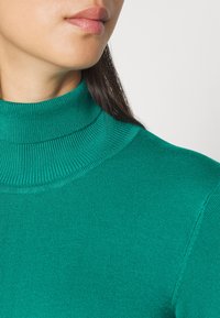Pull en col montant turquoise avec une texture côtelée, un design ajusté et une finition lisse. L'encolure est haute et épouse étroitement la peau.