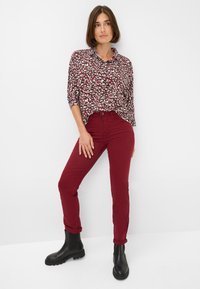 Camicia a stampa floreale color bordeaux, vestibilità ampia con maniche arrotolate, abbinata a pantaloni bordeaux aderenti e stivali neri alla caviglia con suola spessa.