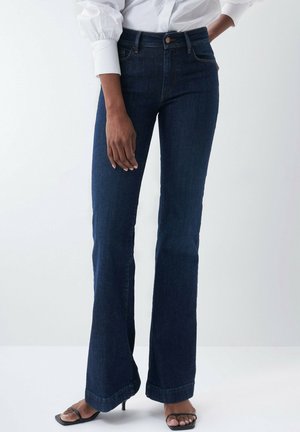 Kobieta ubrana w ciemnoniebieskie dżinsy bootcut, białą bluzkę i czarne sandały na obcasie z paskami, stojąca na tle jasnego, jednolitego tła.