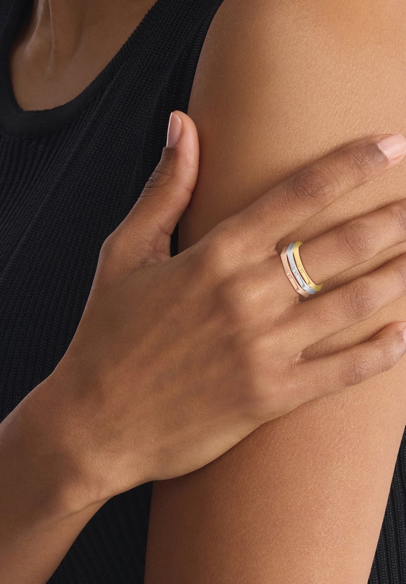 Conjunto de tres anillos apilados en tonos dorado, plateado y rosado, que presentan acabados lisos y bandas de color distintas. Mano reposando sobre un brazo.