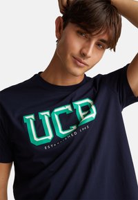 Tmavě modré tričko s krátkým rukávem vyrobené z bavlny, zdobeno nápisem "UCD" v zelenobílém písmu a s nápisem "Založeno 1965" pod ním.